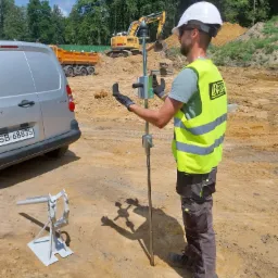 Pracownik w kasku i kamizelce odblaskowej obsługuje urządzenie do badania gruntu na placu budowy; w tle koparka i wywrotka.