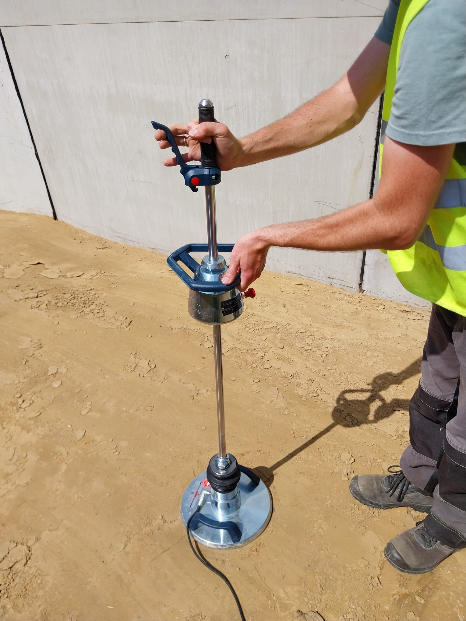 Pracownik w kamizelce odblaskowej używa sondy dynamicznej do badania gruntu na placu budowy, widoczny betonowy mur w tle.