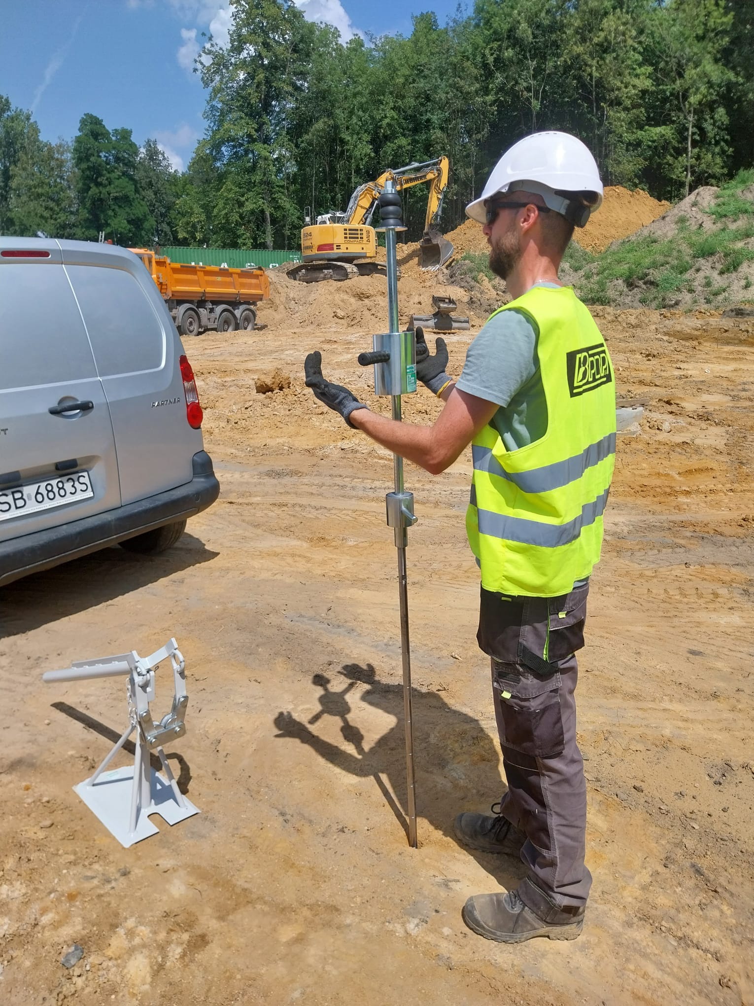 Pracownik w kasku i kamizelce odblaskowej obsługuje urządzenie do badania gruntu na placu budowy; w tle koparka i wywrotka.