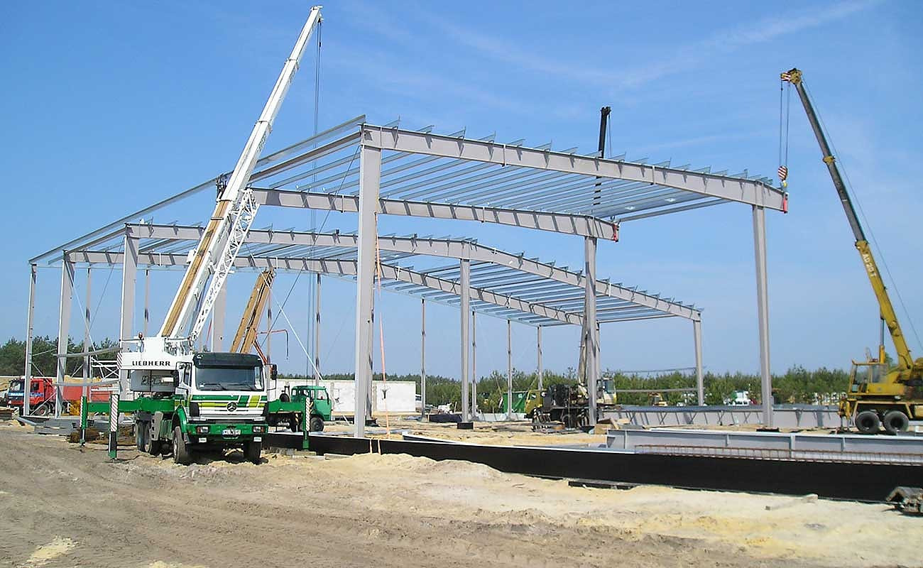 Montaż stalowej konstrukcji hali magazynowej, widoczne dźwigi Liebherr i żółty dźwig kołowy, elementy konstrukcyjne na fundamencie, w tle zieleń.