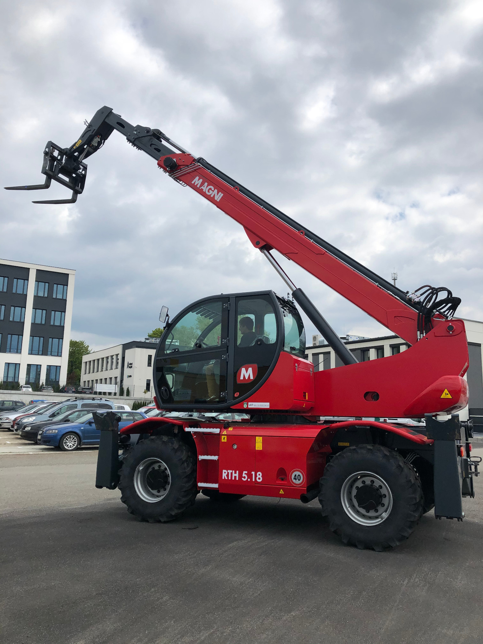 Czerwony, teleskopowy wózek widłowy Magni RTH 5.18 z podniesionym wysięgnikiem na tle parkingu i budynków biurowych w pochmurny dzień.