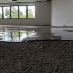 Wylewka samopoziomująca na podłodze w surowym stanie, odbijająca światło z okna, widoczne ściany w trakcie wykończenia.