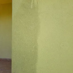 Fragment elewacji budynku w odcieniach żółci, z widoczną różnicą w czystości powierzchni po częściowym umyciu. Dolna część ściany wykończona czerwoną cegłą.