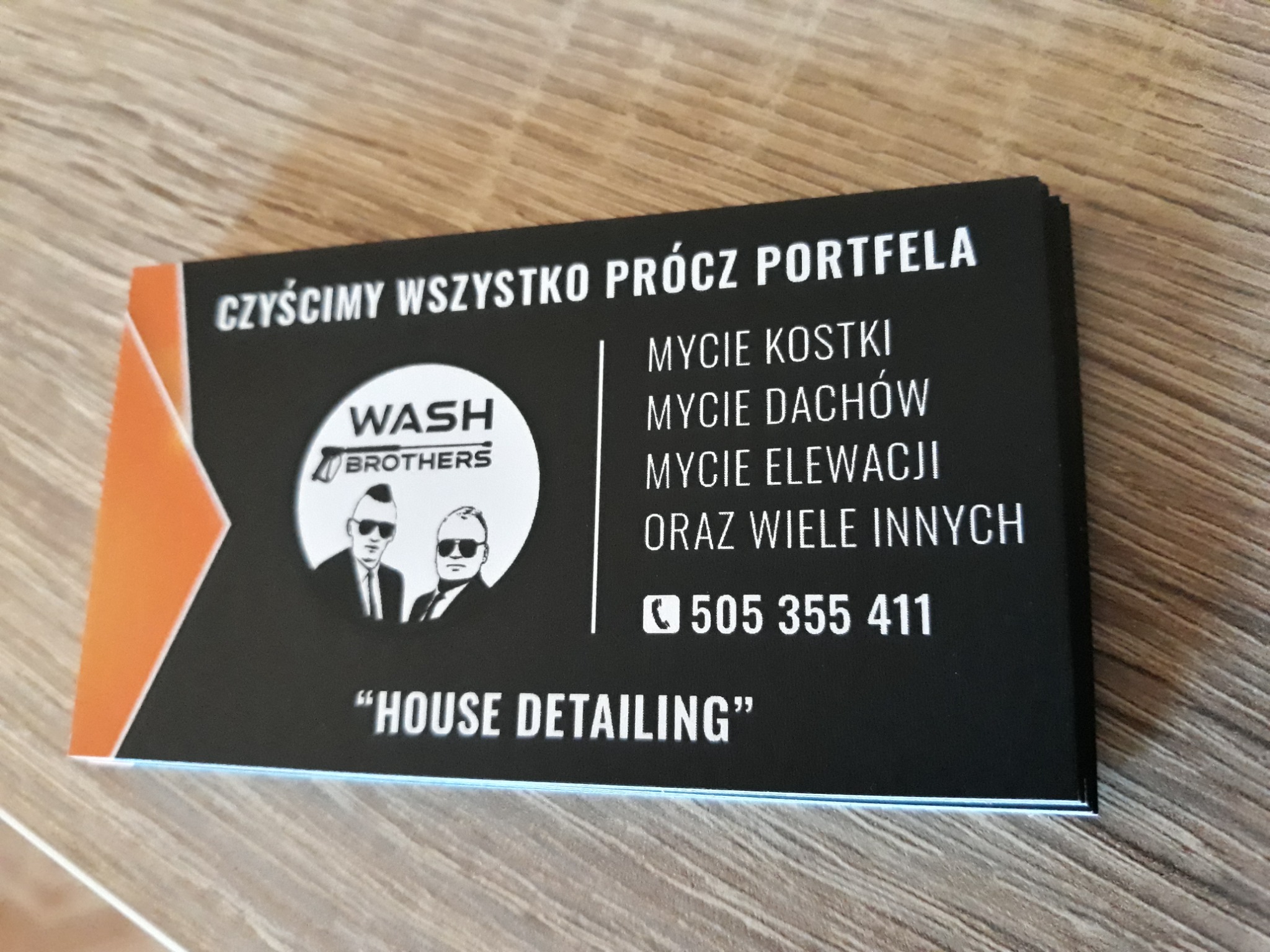 Wizytówka firmy 'Wash Brothers' oferującej usługi czyszczenia, w tym kostki brukowej, dachów i elewacji, z numerem telefonu 505 355 411, leżąca na drewnianym blacie.