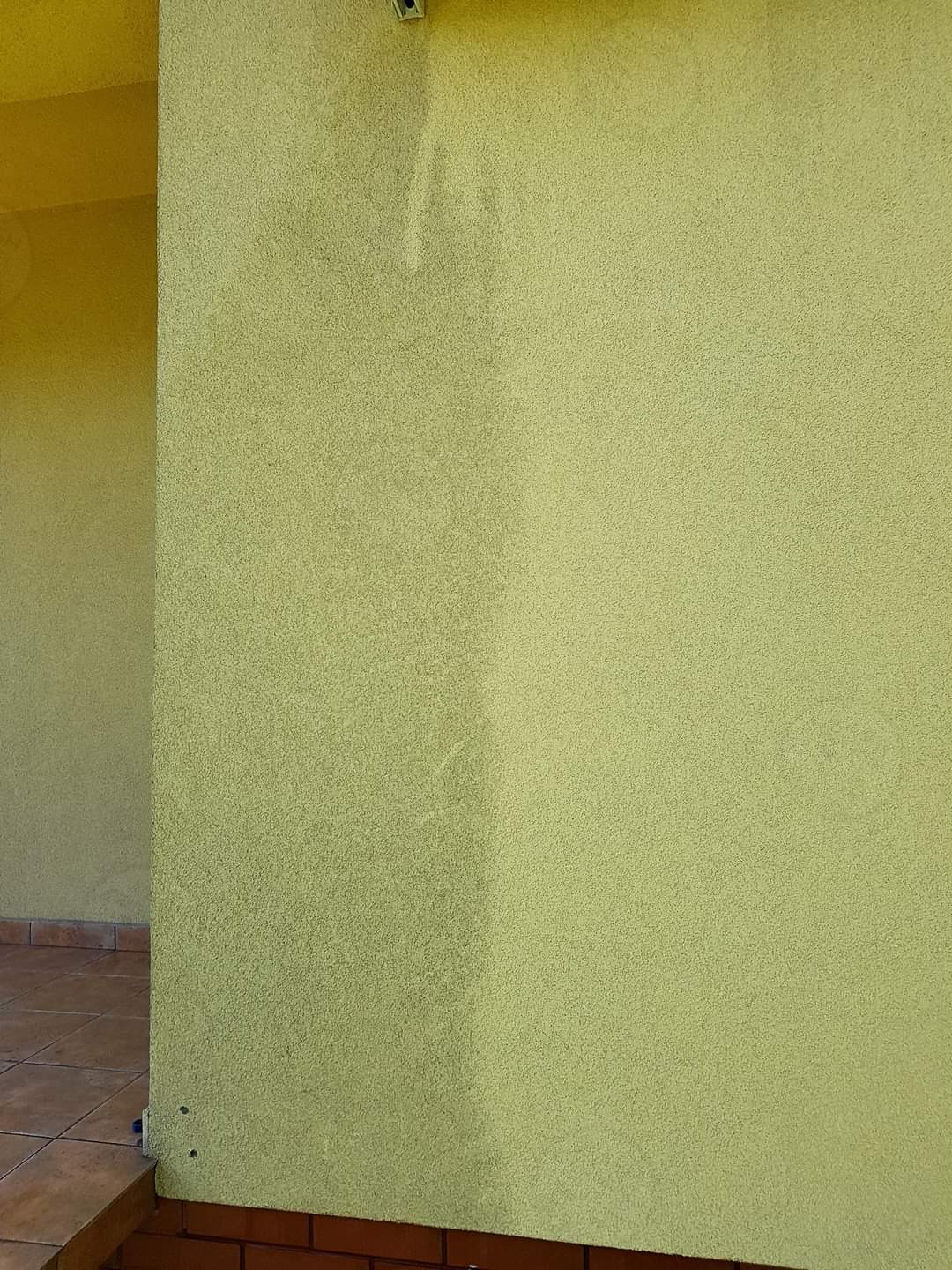 Fragment elewacji budynku w odcieniach żółci, z widoczną różnicą w czystości powierzchni po częściowym umyciu. Dolna część ściany wykończona czerwoną cegłą.