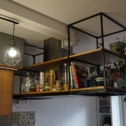 Metalowa konstrukcja regału kuchennego podwieszona pod sufitem, z drewnianymi półkami wypełnionymi książkami, przyprawami i roślinami doniczkowymi, oświetlona przez lampy wiszące w stylu...