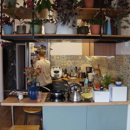 Marcin Birski - Wnętrze kuchni z wyspą kuchenną, na której znajdują się garnki, czajnik, kwiaty w wazonie, a nad nią półki z roślinami doniczkowymi; osoba stoi przy otwartej lodówce.