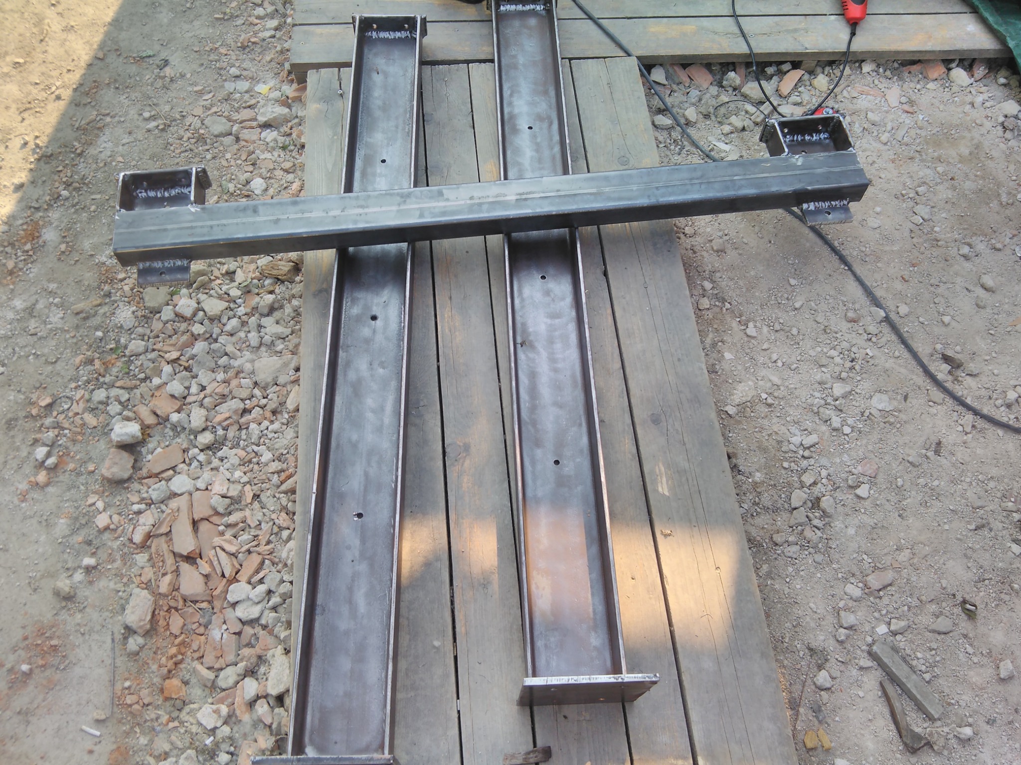Metalowe profile, dwie pionowe kolumny i jeden poziomy element, z widocznymi śladami łączenia, ułożone na drewnianej powierzchni na zewnątrz.