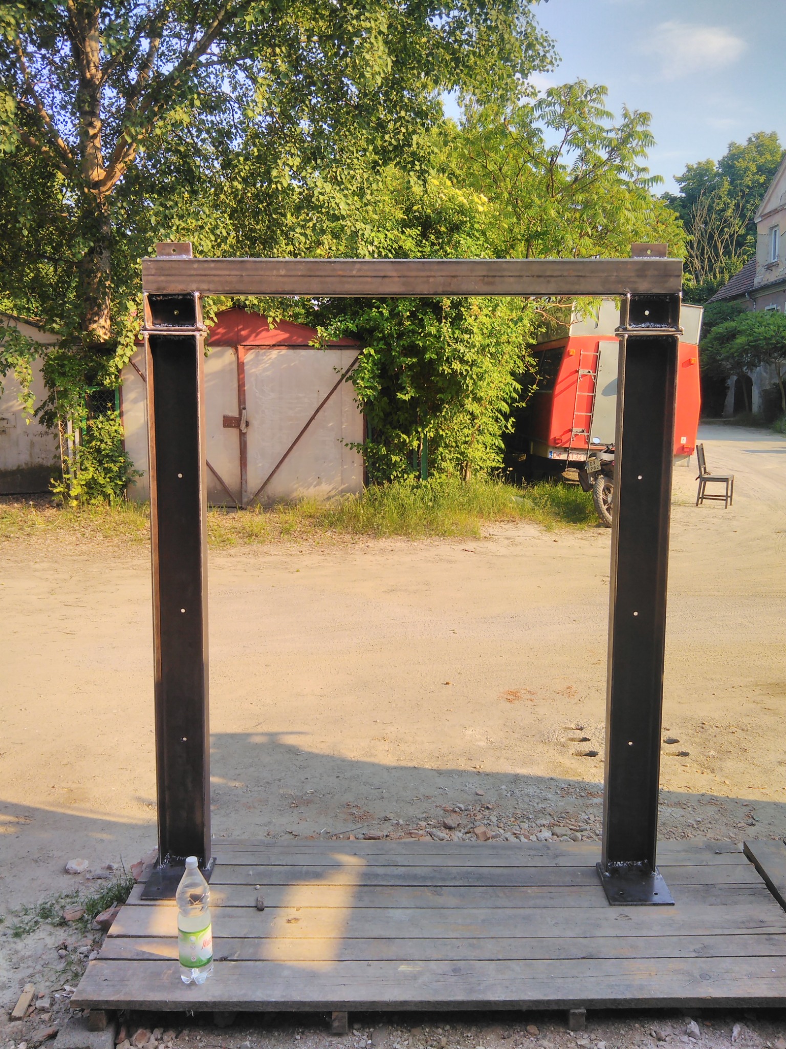 Metalowa rama konstrukcyjna z profili zamkniętych, ustawiona na drewnianym podeście, butelka wody u podstawy, w tle zabudowania gospodarcze i zieleń.