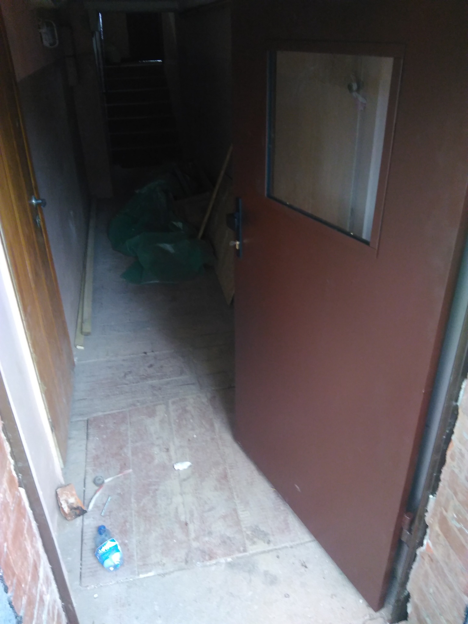 Otwarta brązowa stalowa brama z małym okienkiem, prowadząca do ciemnego korytarza ze schodami. Na podłodze leży butelka wody i drobne śmieci.