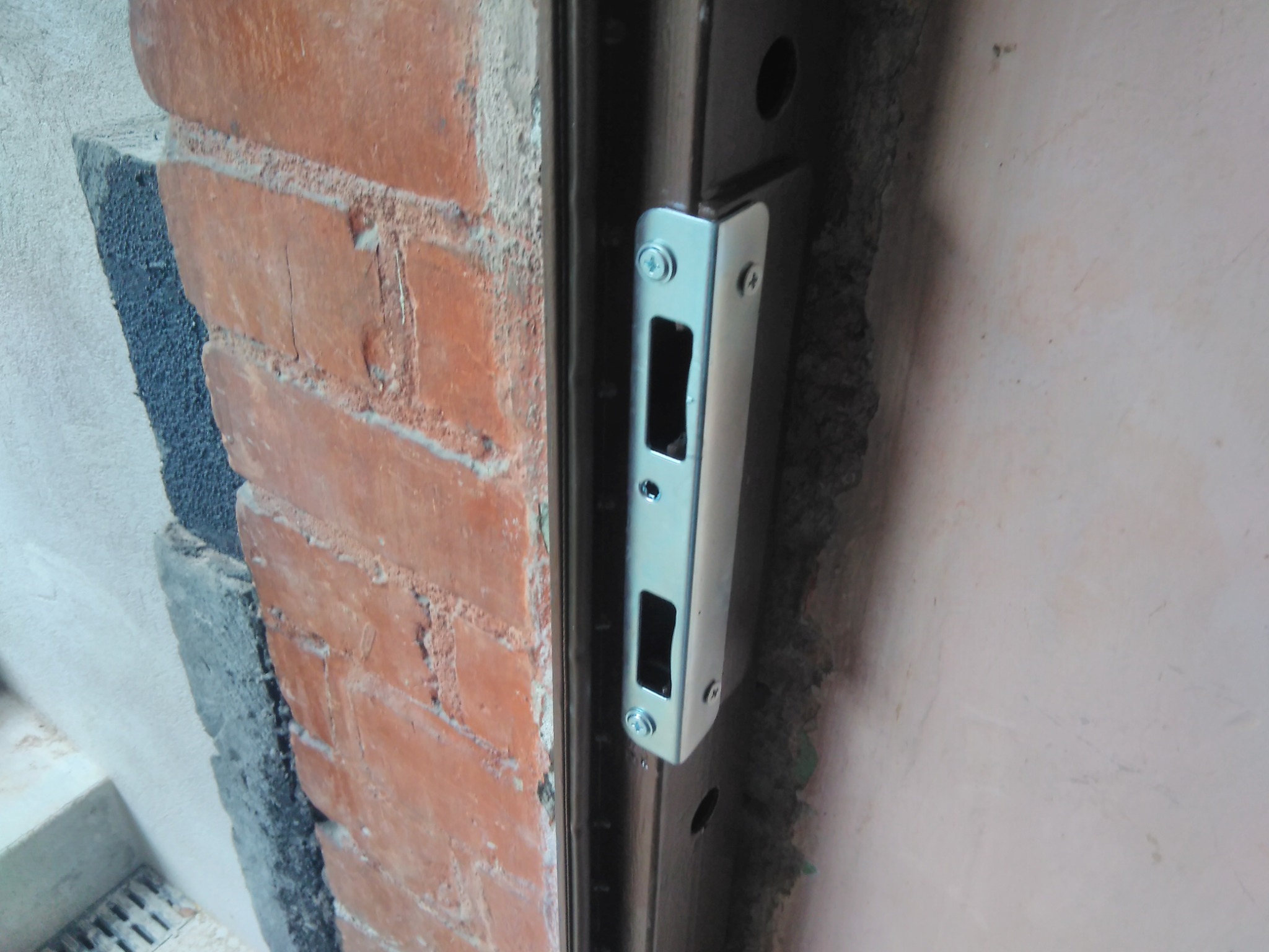 Montaż stalowej futryny drzwiowej z zamkiem w murze z czerwonej cegły, widoczne śruby mocujące i fragment ściany z tynkiem.