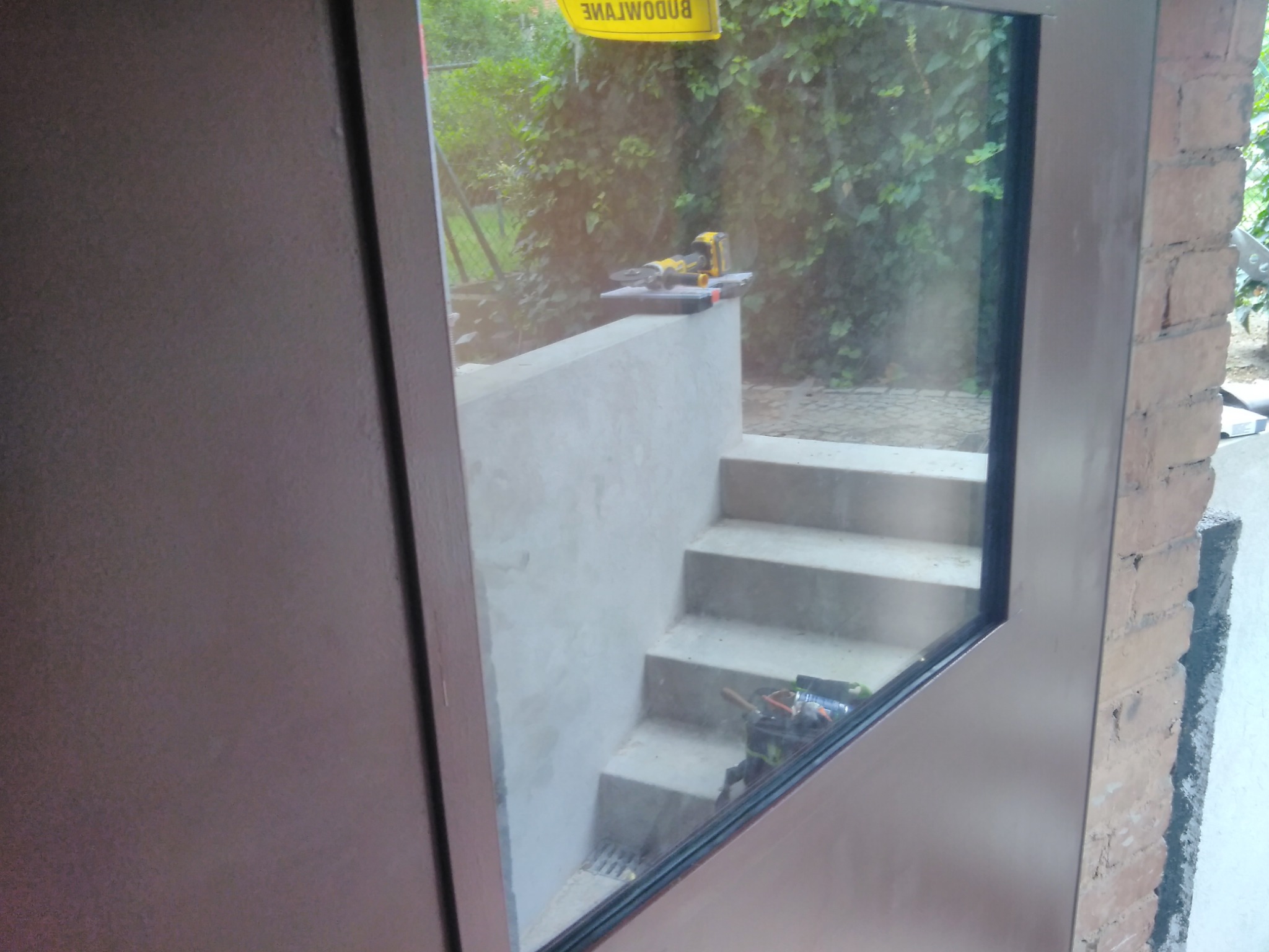 Widok przez okno na betonowe schody piwniczne z narzędziami na stopniach i na górnej krawędzi, ceglany mur po prawej stronie.