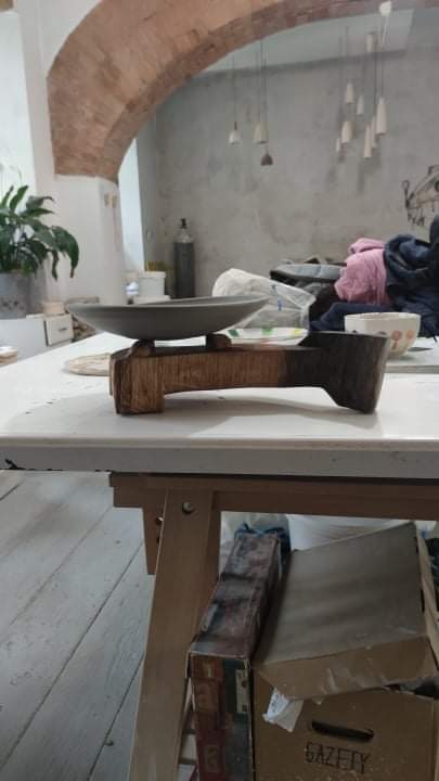 Szara, ceramiczna misa na drewnianej podstawie w pracowni artystycznej z ceglanym łukiem i wiszącymi lampami w tle.