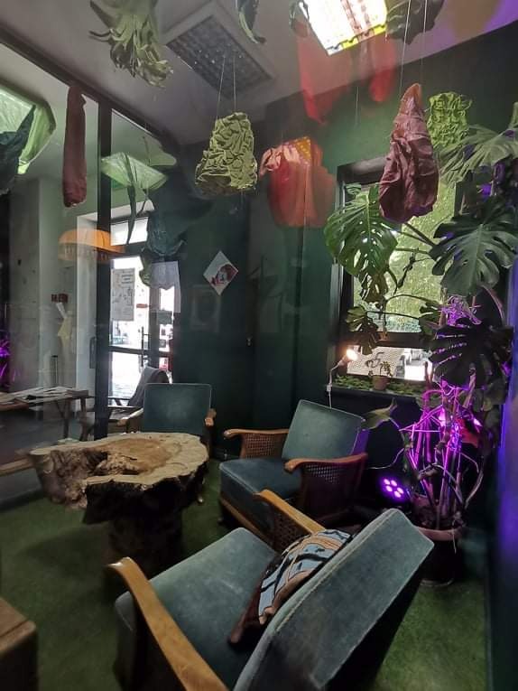 Wnętrze kawiarni z surowym drewnianym stolikiem, czterema tapicerowanymi fotelami w odcieniach zieleni i dekoracjami wiszącymi pod sufitem, oświetlone fioletowym światłem.