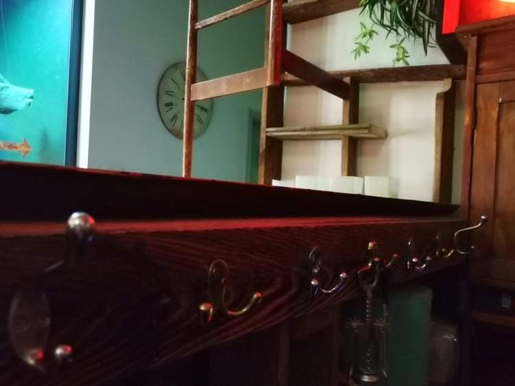 Drewniana półka z haczykami w stylu rustykalnym, widoczny zegar na ścianie w tle oraz elementy dekoracyjne, w tym roślina doniczkowa.