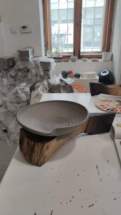 Szary, ceramiczny talerz z wgłębieniem na drewnianej podstawie, w tle okno z kratami i stosy zapakowanej gliny.