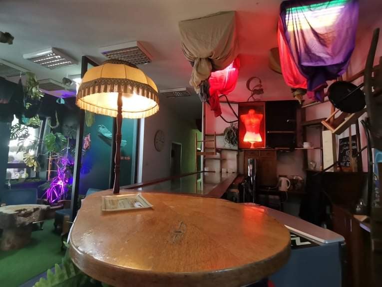 Wnętrze artystycznej pracowni z drewnianym blatem stołu na pierwszym planie, lampą z frędzlami, akwarium z roślinami i podwieszonymi materiałami w roli oświetlenia.