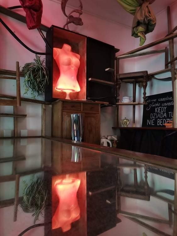 Podświetlony, różowy manekin damski w drewnianej skrzyni, odbijający się w szklanym blacie, otoczony eklektycznym wystrojem wnętrza z drewnianymi elementami, roślinami i tablicą z napisem.
