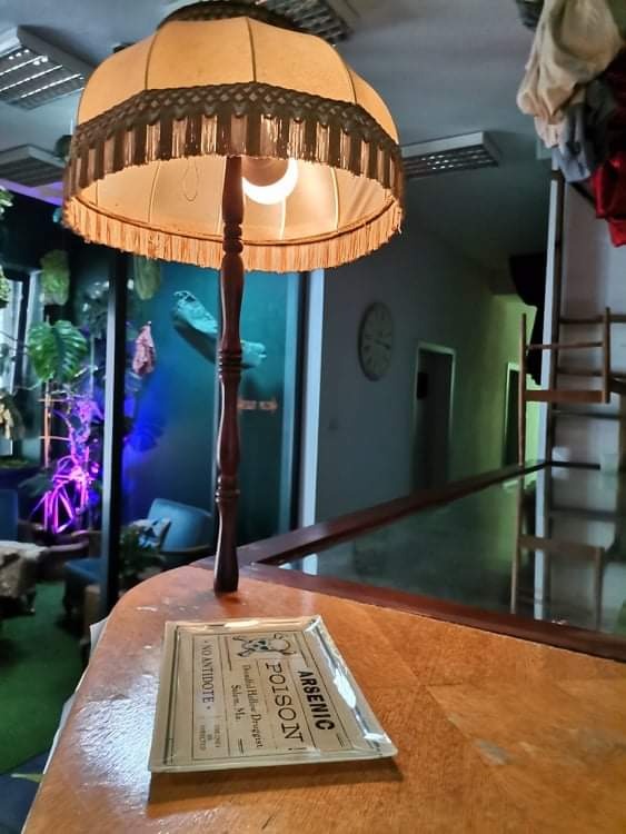 Retro lampa podłogowa z abażurem z frędzlami, stojąca na drewnianym blacie, obok szklana taca z napisem 'Arsenic Poison', w tle niebieskie światło i elementy wyposażenia wnętrza.