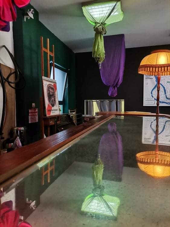 Ekscentryczne wnętrze z elementami artystycznymi: drewniany stół barowy, lampa z mapą w tle, zielone i fioletowe tkaniny zwisające z sufitu, obraz na sztaludze przy ścianie w ciemnych odcieniach...