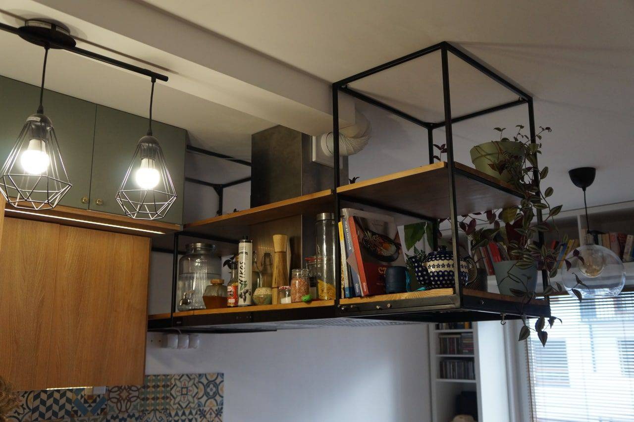 Metalowa konstrukcja regału kuchennego podwieszona pod sufitem, z drewnianymi półkami wypełnionymi książkami, przyprawami i roślinami doniczkowymi, oświetlona przez lampy wiszące w stylu...