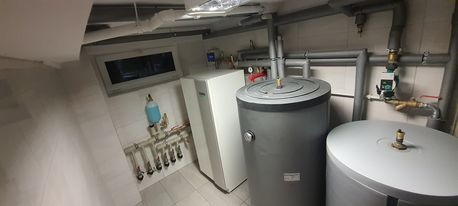 Wnętrze kotłowni z zasobnikami ciepłej wody i pompą ciepła, widoczne liczne rury i zawory, białe płytki na ścianach i podłodze, małe okno w tle.