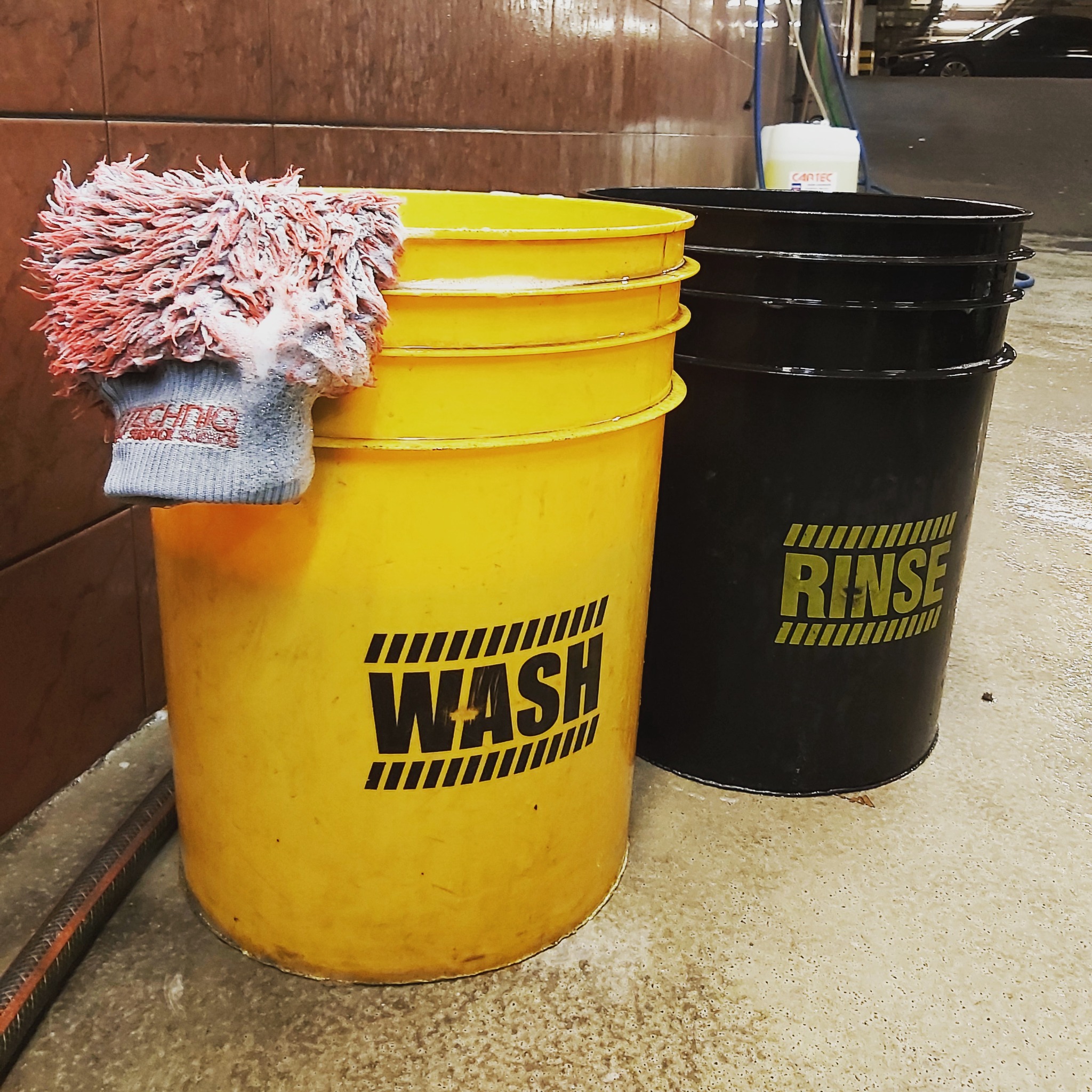 Dwa plastikowe wiadra: żółte z napisem 'WASH' i czarne z napisem 'RINSE', obok leży mokra rękawica do mycia, w tle widoczny wjazd do garażu.