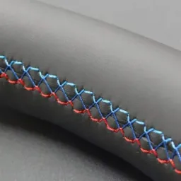 Fragment obszytej skórą kierownicy samochodowej z dekoracyjnym, ręcznym szwem w kolorach niebieskim i czerwonym.