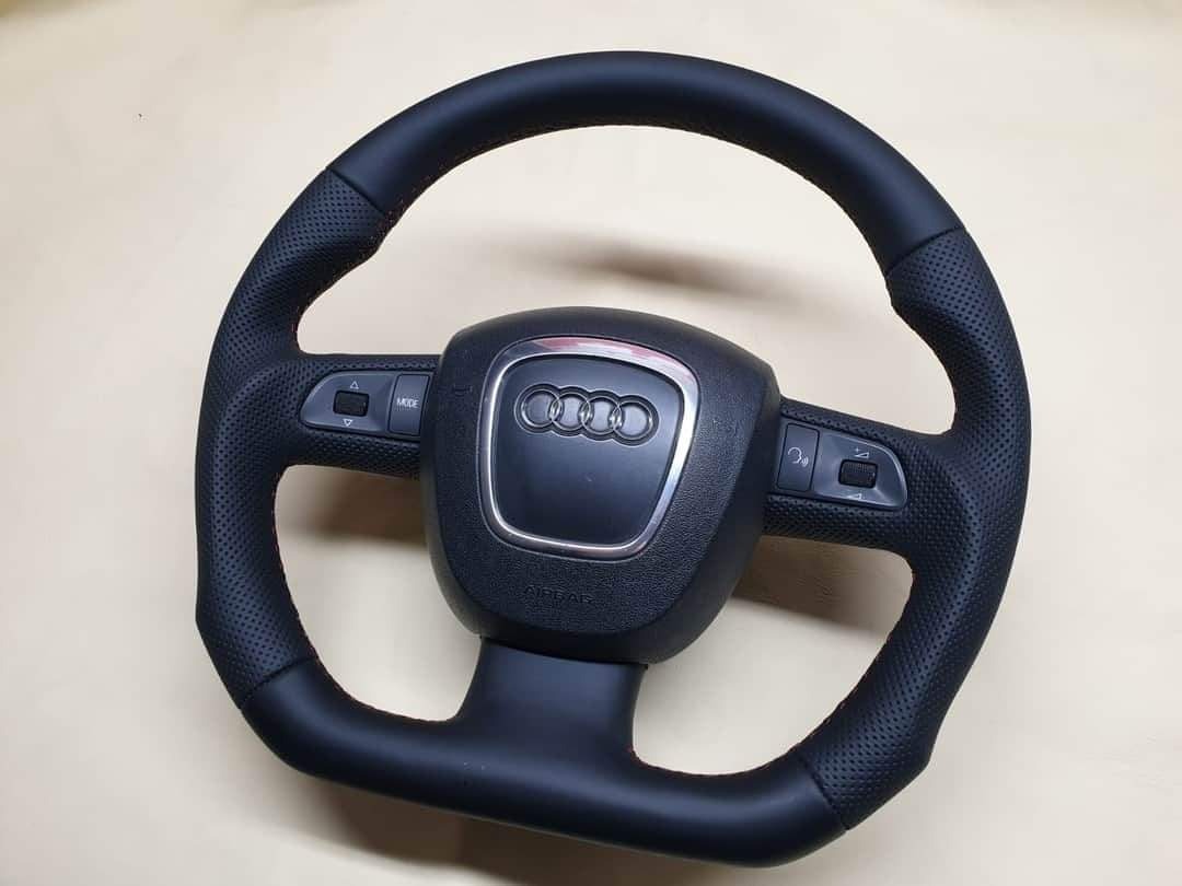 Obszyta czarną skórą kierownica Audi z perforacją i czerwonymi przeszyciami, widoczne logo marki.
