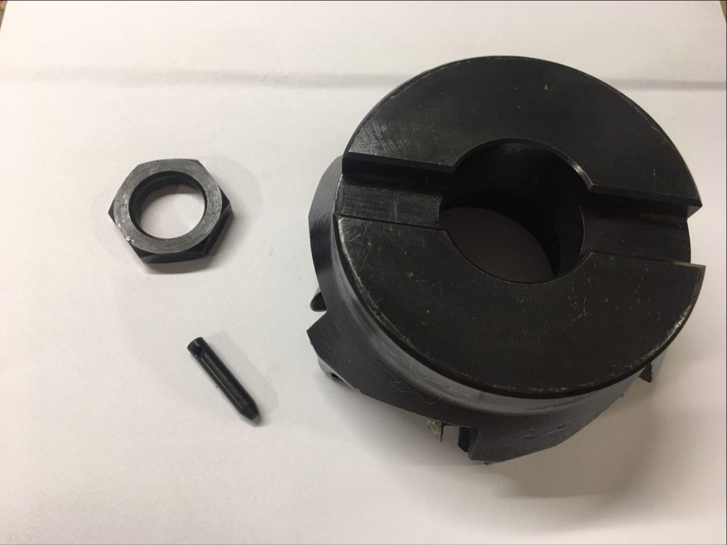 Metalowa tuleja zaciskowa, nakrętka i kołek na białym tle, widoczne ślady obróbki mechanicznej.