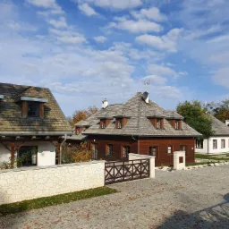 Kompleks budynków w stylu dworkowym z drewnianymi dachami krytymi gontem, otoczony brukowanym dziedzińcem i niskimi murkami z kamienia łupanego.