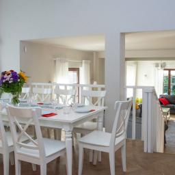 Wzgórze Zamkowe Sp. z o.o. - Biały stół z nakryciem i bukietem kwiatów w jasnym wnętrzu domu, widok na salon z szarą sofą, drewniana podłoga w jodełkę.