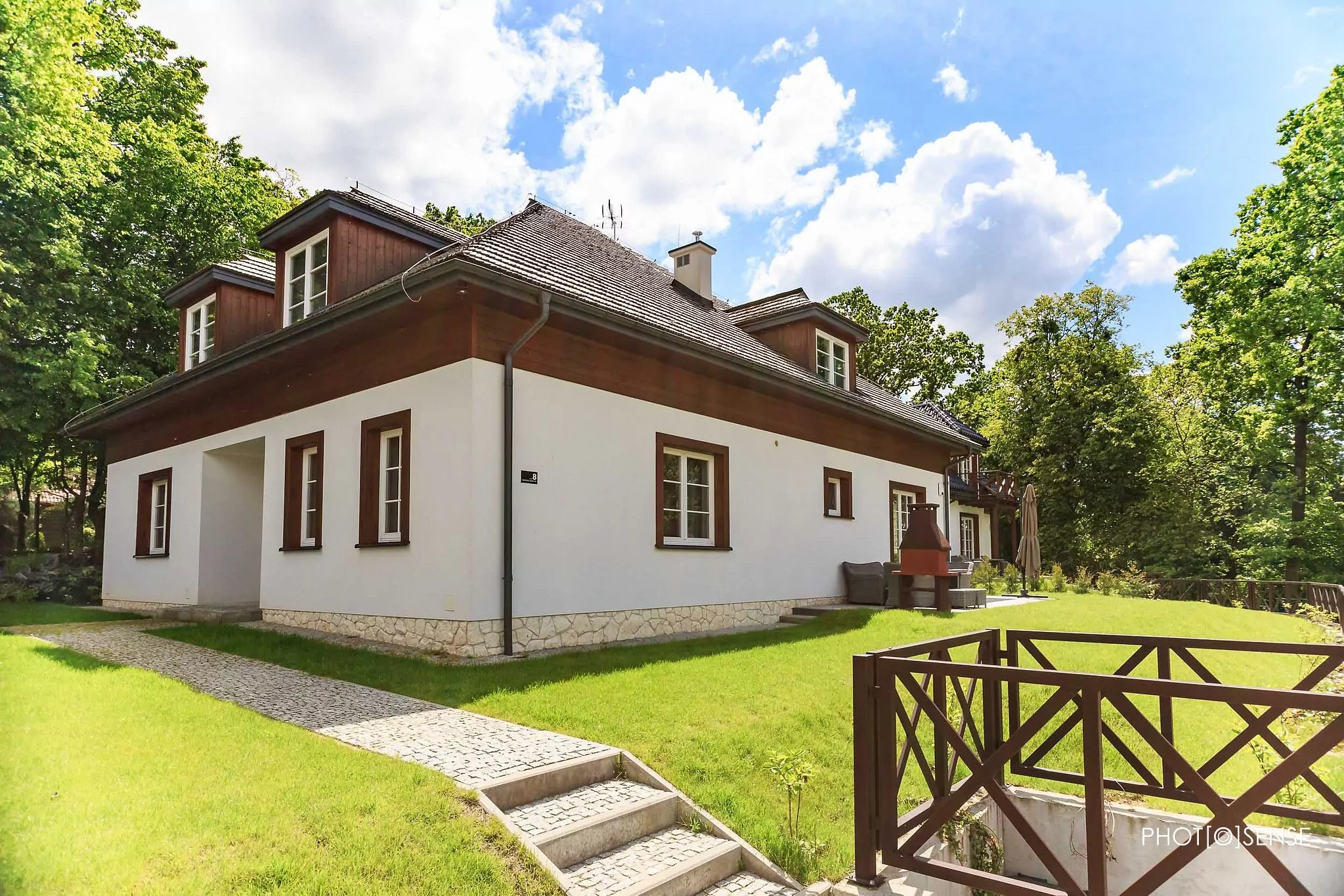 Biały dom z brązowymi elementami wykończenia, widoczny z zewnątrz, z kamienną ścieżką prowadzącą do wejścia i zadbanym trawnikiem, otoczony zielenią drzew, z numerem 8 na elewacji.