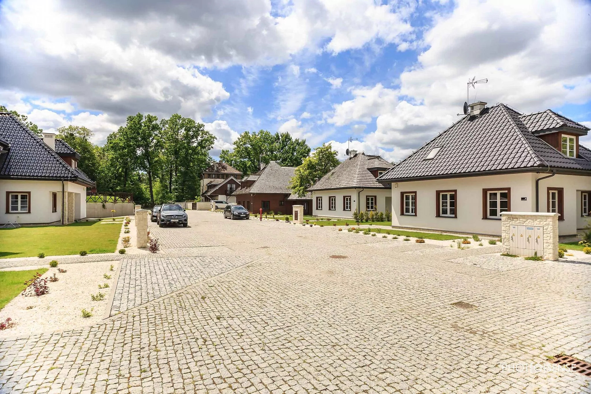 Kompleks budynków z ciemnymi dachami i białymi ścianami, wybrukowany dziedziniec z samochodami w tle, błękitne niebo z chmurami.