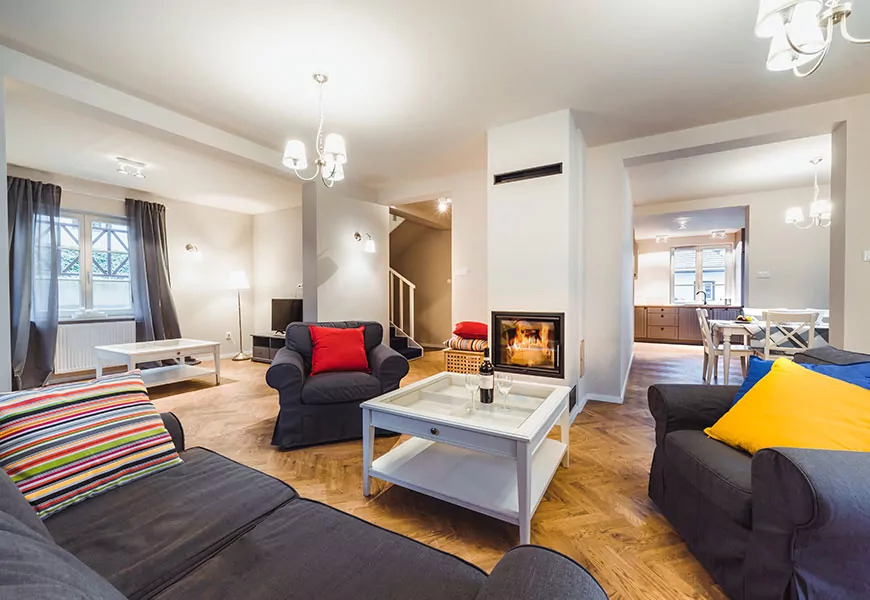 Jasne wnętrze pokoju dziennego z kominkiem, sofami i widokiem na jadalnię, na stoliku dwie lampki wina.