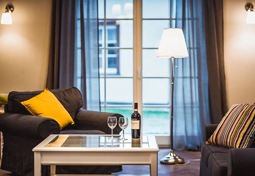 Salon z dwoma ciemnymi fotelami, butelką wina i dwoma kieliszkami na białym stoliku kawowym, widok przez okno na zewnątrz, lampy ścienne i podłogowa.