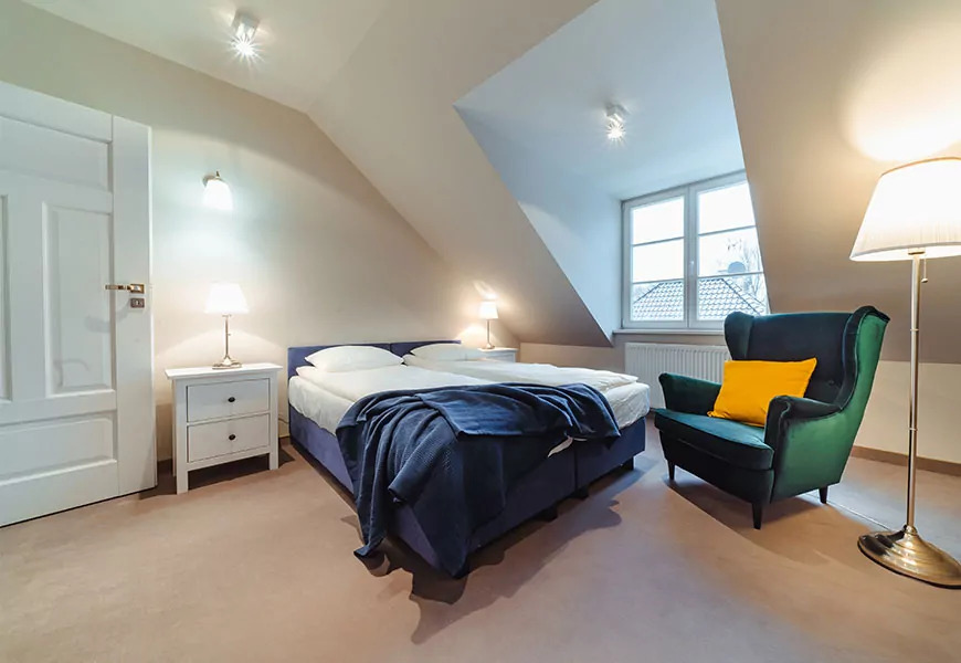 Pokój w hotelu SPA na poddaszu z dwoma łóżkami, granatową narzutą, zielonym fotelem z żółtą poduszką i lampą podłogową.