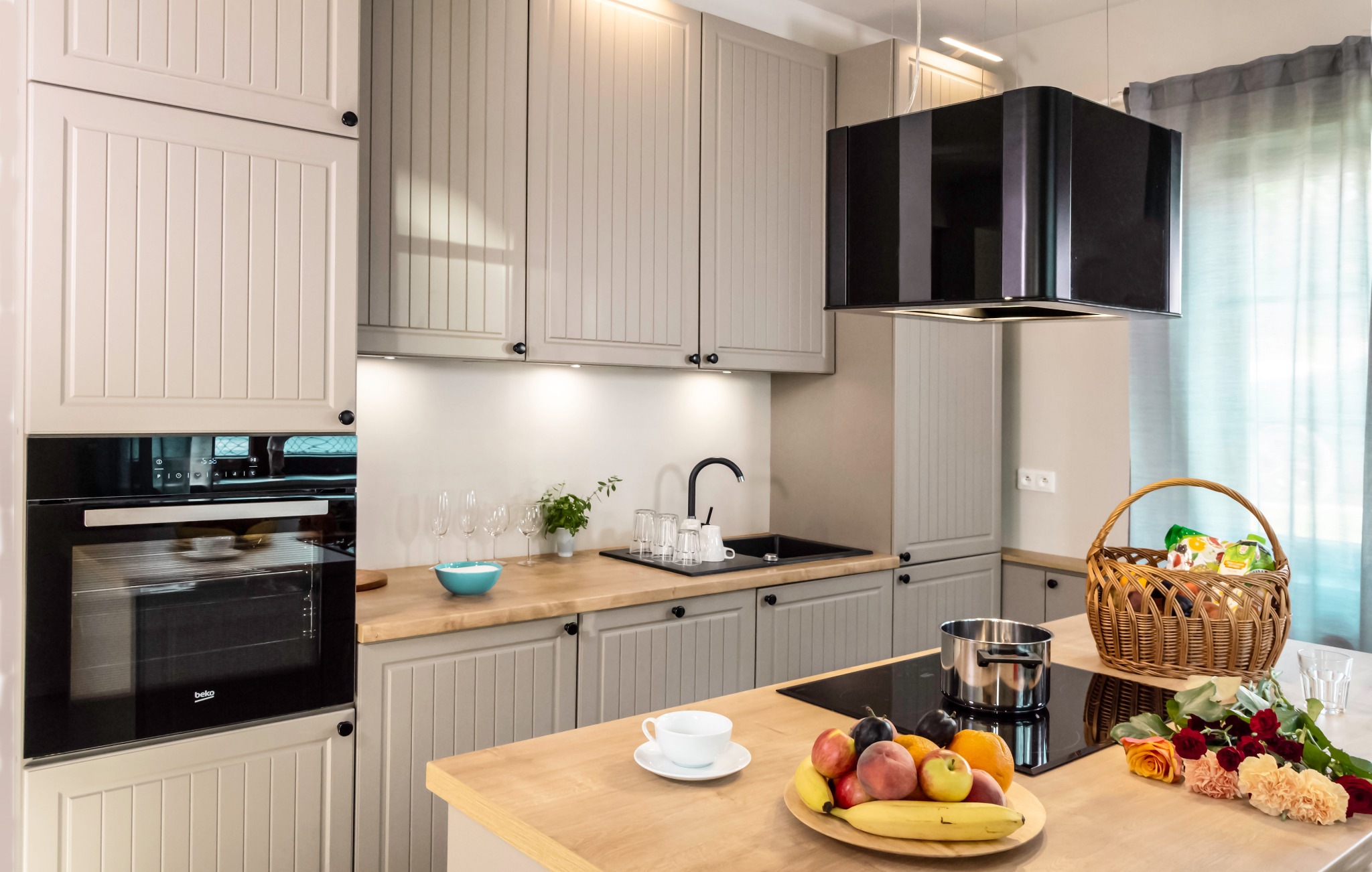 Jasna kuchnia z drewnianym blatem, czarną armaturą i płytą indukcyjną, na blacie owoce, kwiaty, filiżanka, w koszu produkty spożywcze