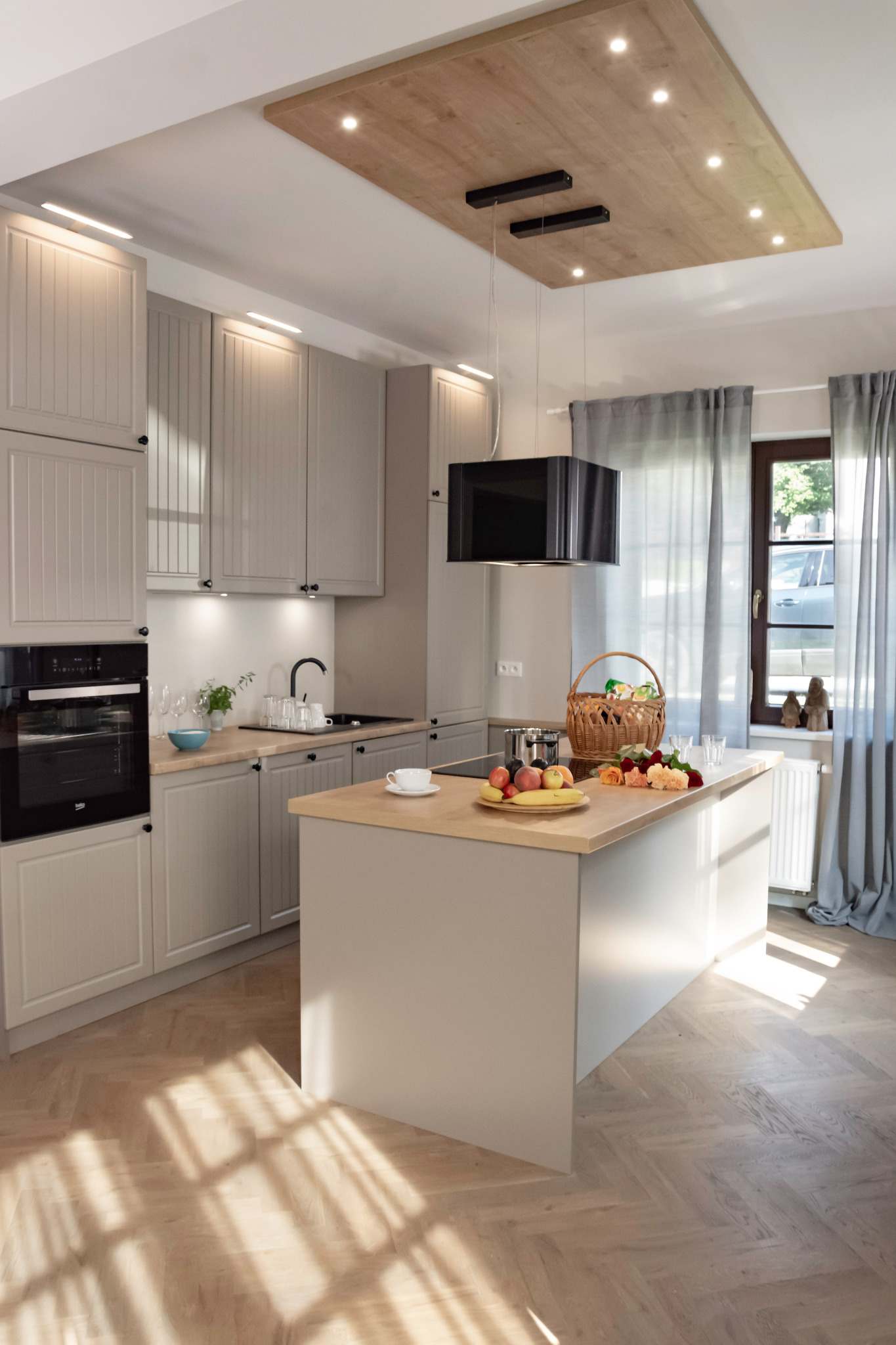 Jasna kuchnia w stylu skandynawskim z drewnianym blatem wyspy, owocami, kwiatami i koszem piknikowym na wyspie, z oknem w tle i parkietem w jodełkę na podłodze.