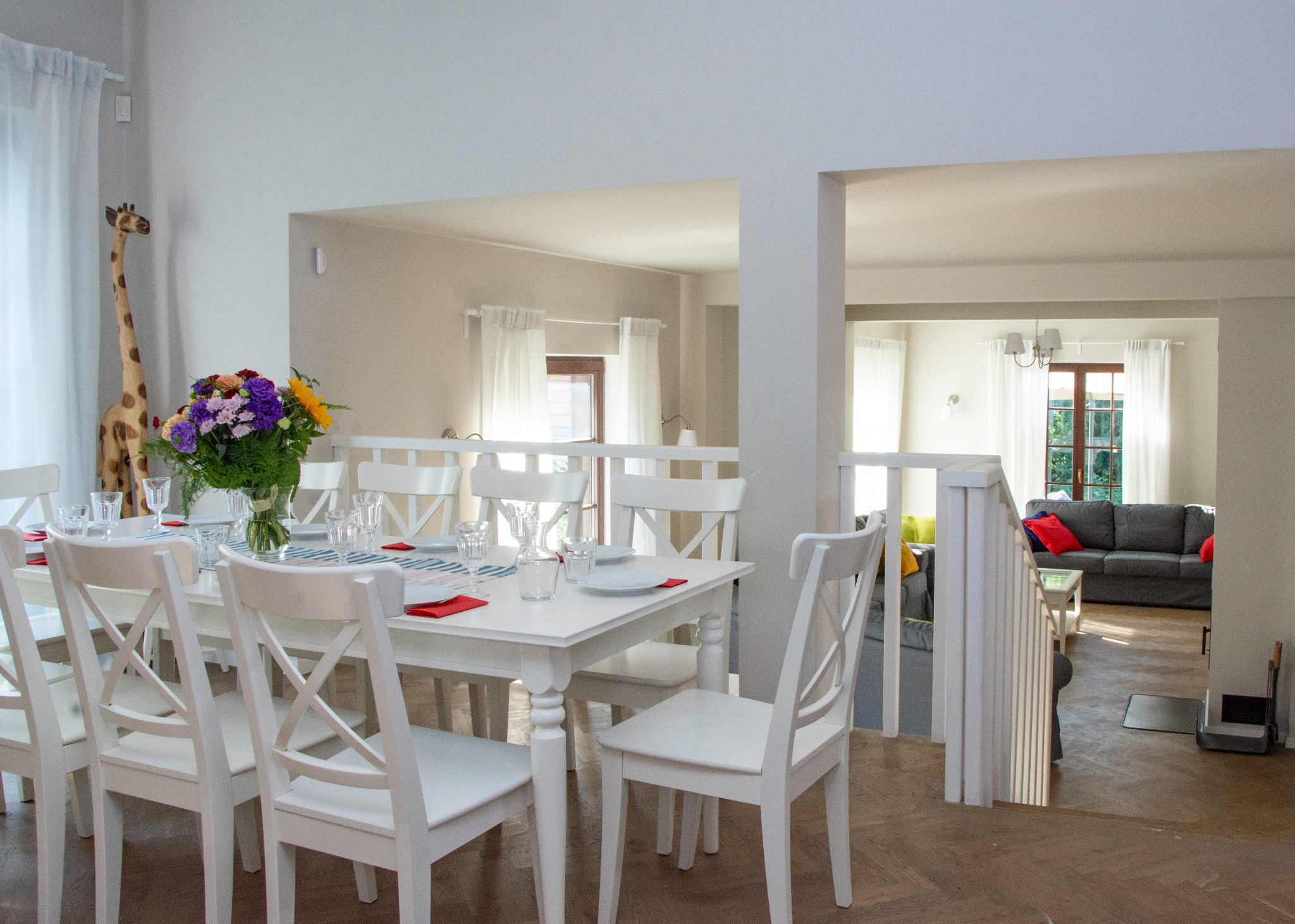 Biały stół z nakryciem i bukietem kwiatów w jasnym wnętrzu domu, widok na salon z szarą sofą, drewniana podłoga w jodełkę.