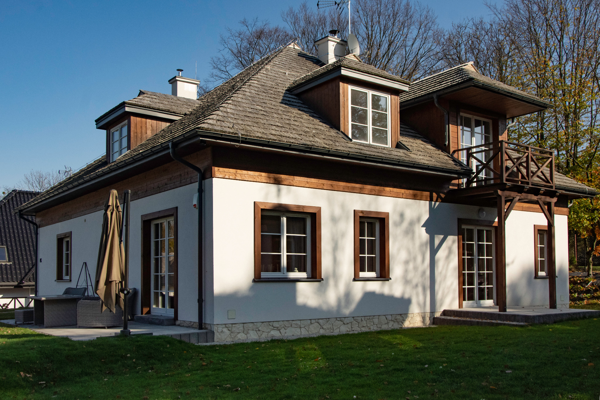 Dwupiętrowy dom z brązowymi drewnianymi obramowaniami okien i dachem z dachówki bitumicznej, z małym balkonem z drewnianą balustradą i zielonym trawnikiem przed budynkiem.