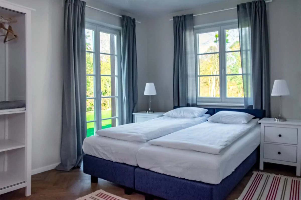 Jasny pokój z dwoma łóżkami pojedynczymi, szafkami nocnymi i widokiem na zieleń za oknem.