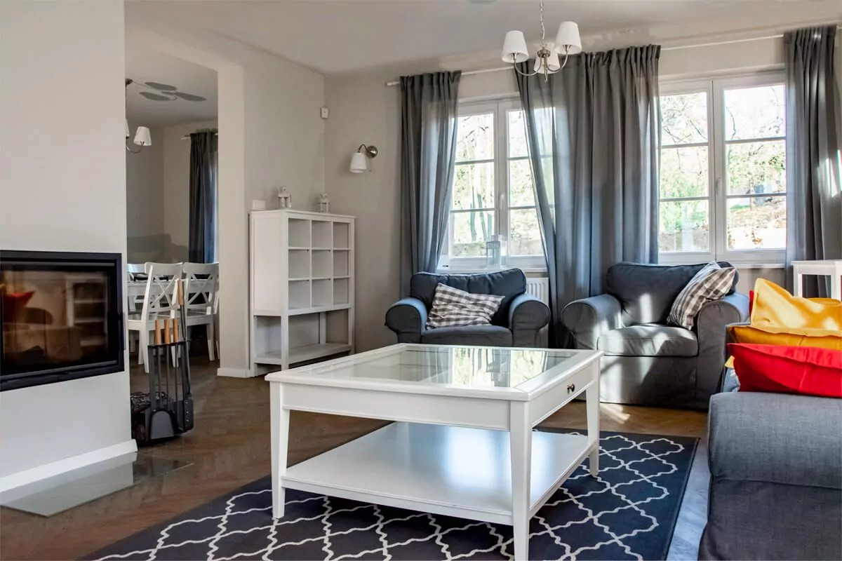 Jasny salon z kominkiem, dwoma szarymi fotelami, białym stolikiem kawowym z szklanym blatem i wzorzystym dywanem na podłodze.