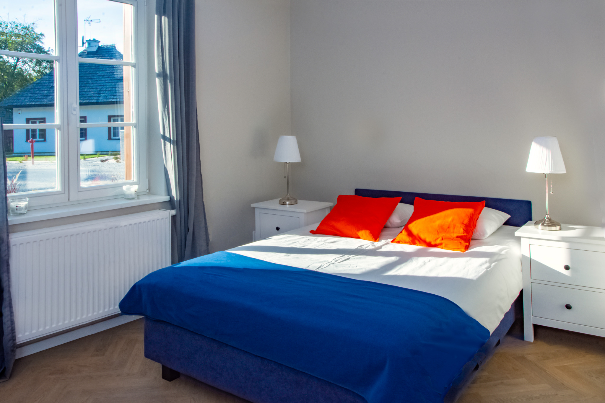 Jasny pokój hotelowy z dużym łóżkiem, pomarańczowymi poduszkami i widokiem na budynek przez okno.