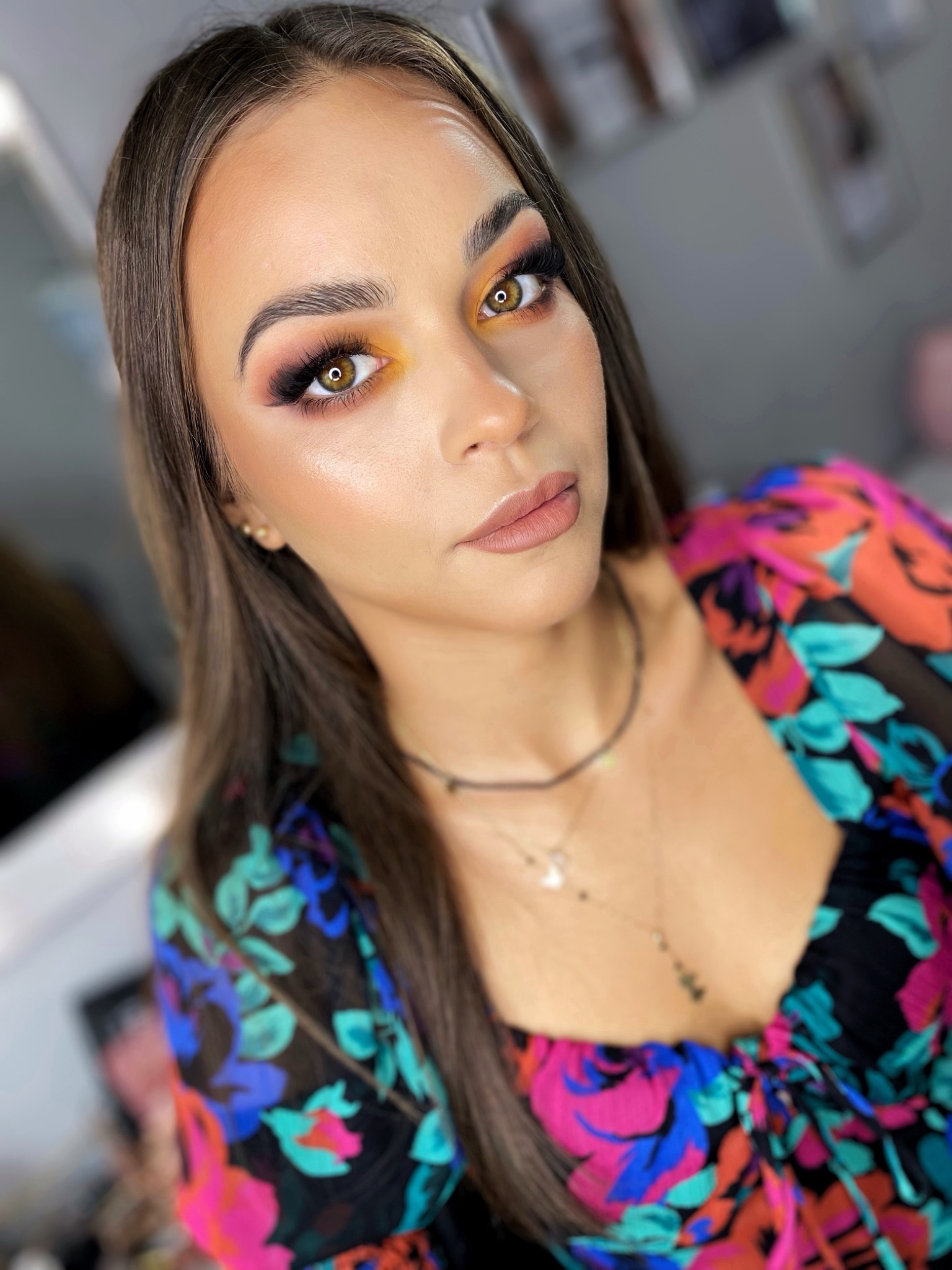 Portret kobiety z profesjonalnie wykonanym, kolorowym makijażem oczu w odcieniach pomarańczy i różu, podkreślonymi brwiami i beżowymi ustami, ubrana w bluzkę w kwiatowy wzór, z delikatnym naszyjnikiem