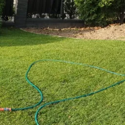 Panoramiczny widok na zadbany trawnik z systemem nawadniania (wąż ogrodowy), elementami brukowanymi i meblami ogrodowymi w tle.