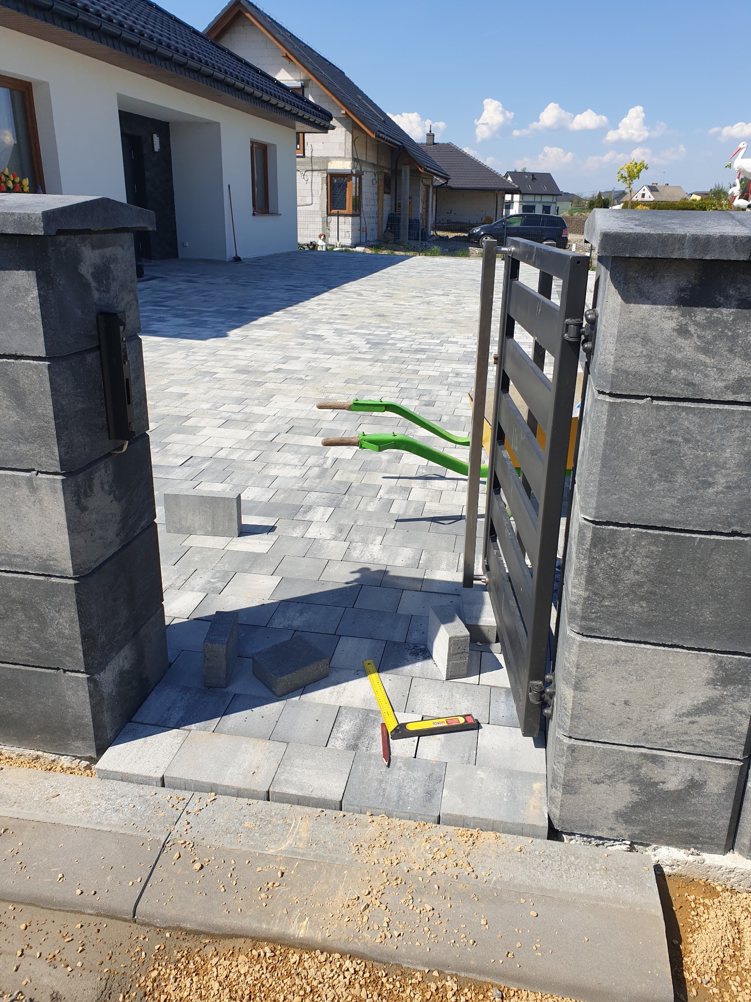 Fragment posesji w trakcie prac brukarskich i montażu bramy: szara kostka brukowa ułożona na podjeździe, czarna brama częściowo zamontowana między dwoma słupkami, poziomica i niedokończone...