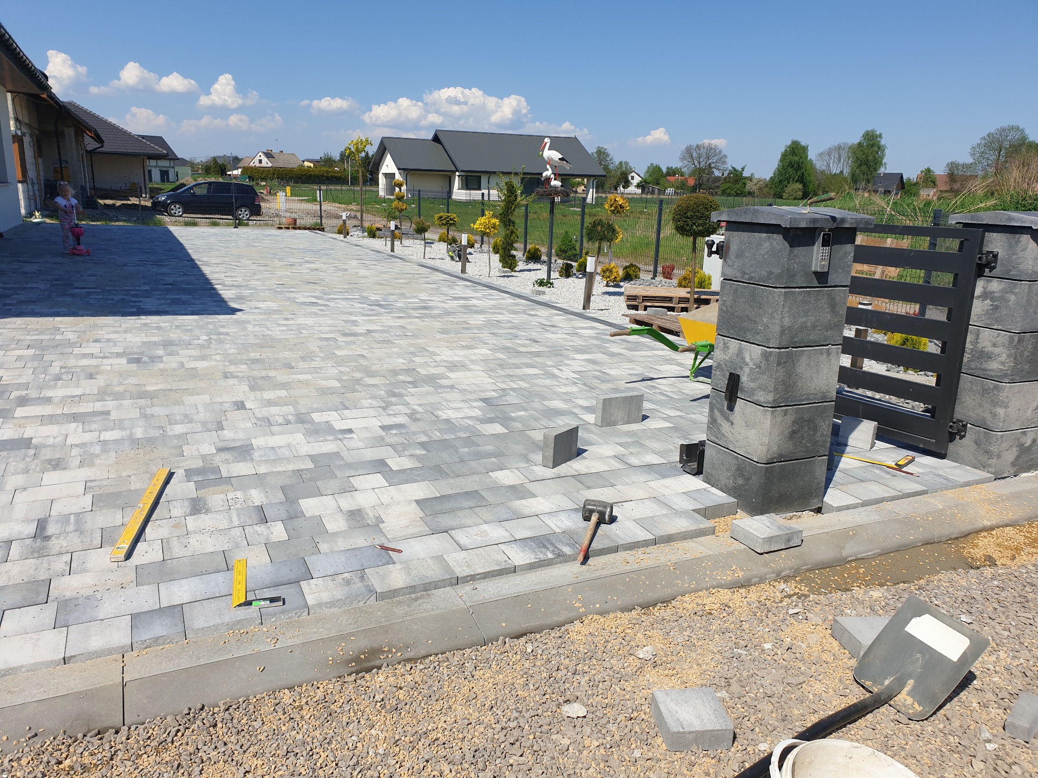 Ułożona kostka brukowa na podjeździe, widoczne narzędzia brukarskie, brama wjazdowa z domofonem i ozdobne rośliny w tle, słoneczny dzień.