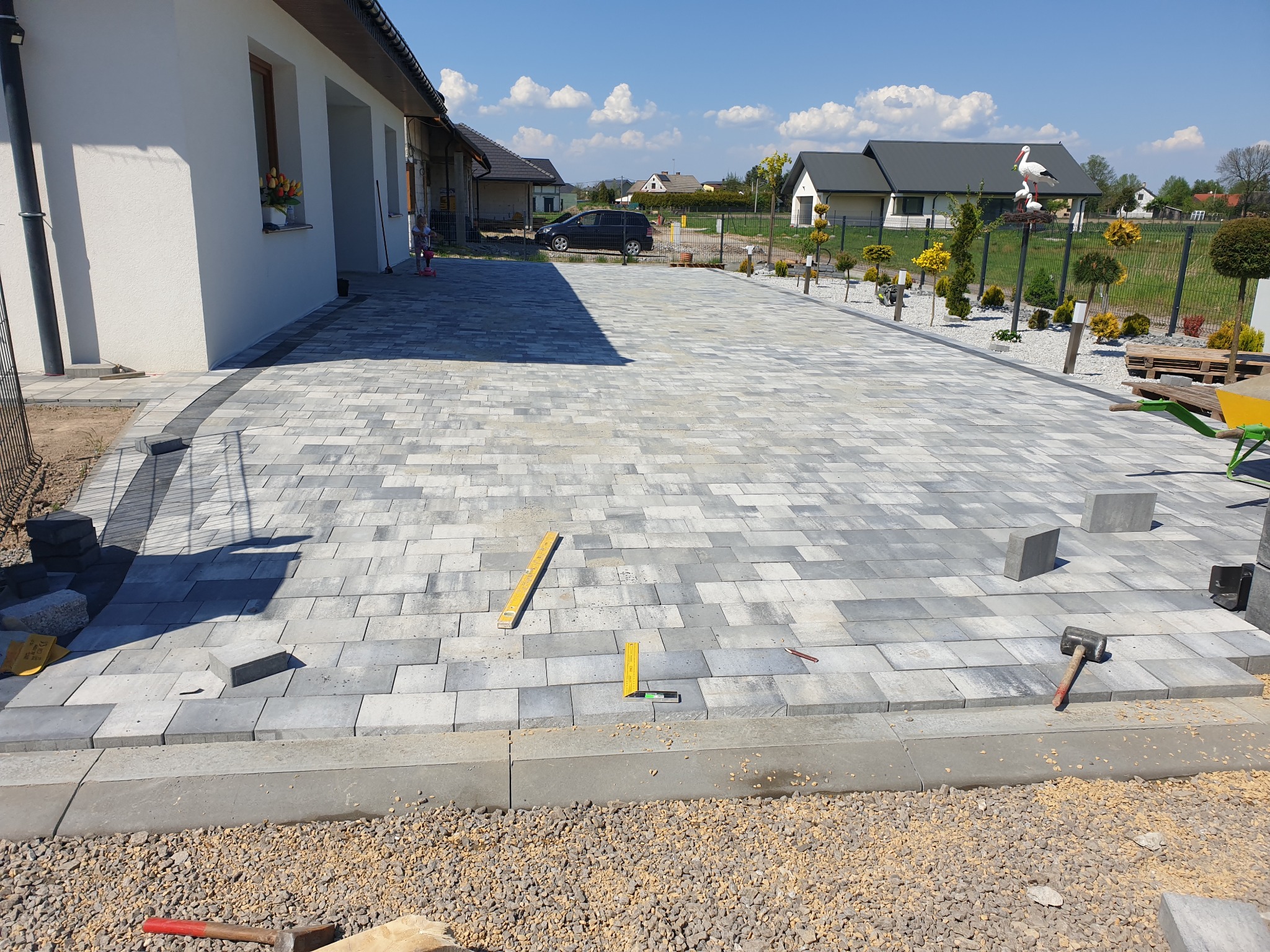 Szara kostka brukowa ułożona na podjeździe przed domem jednorodzinnym, widoczne narzędzia pracy i niezakończony fragment układania.