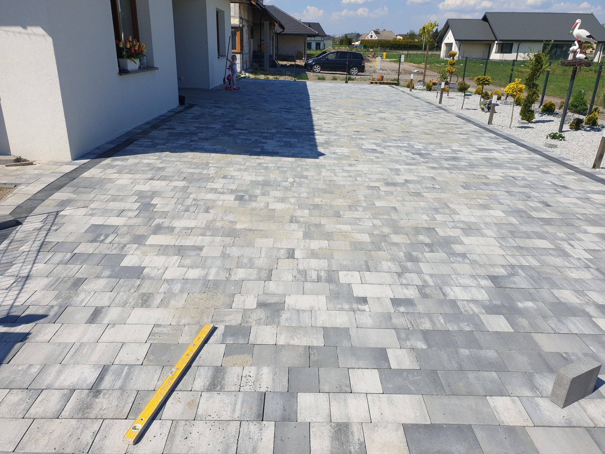 Nowo ułożona, szara kostka brukowa na podjeździe z widocznym poziomem żółtym, budynkiem w tle i elementami dekoracyjnymi w ogrodzie.